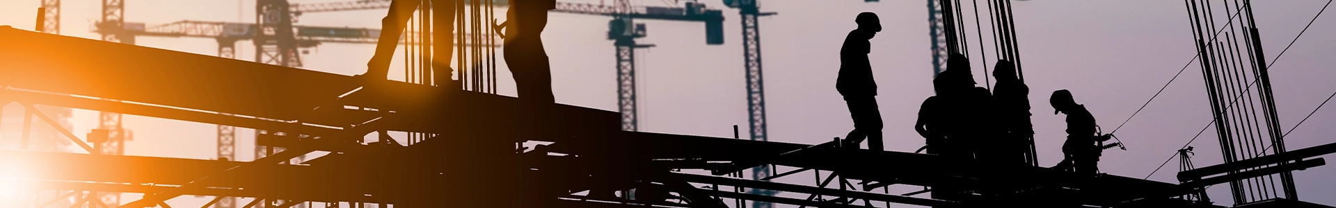 Silhouetted construction workers on a building framework with multiple cranes in the background at sunset.