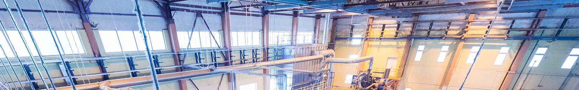Interior view of an industrial manufacturing facility with large metal pipes, overhead structures, and bright window lighting.
