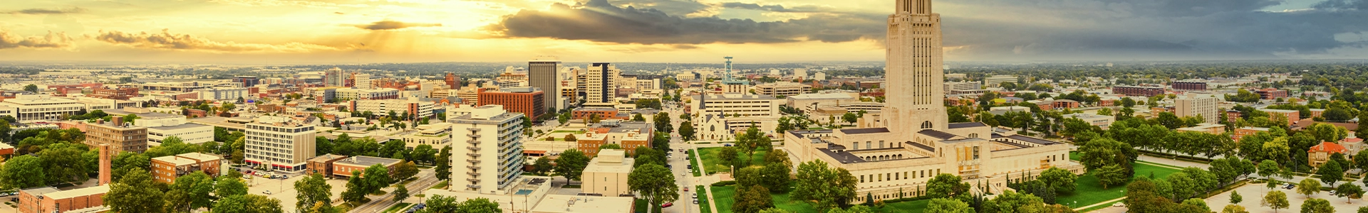 Panoramic view of downtown Lincoln featuring the Nebraska State Capitol, city buildings, and surrounding green spaces at sunset.