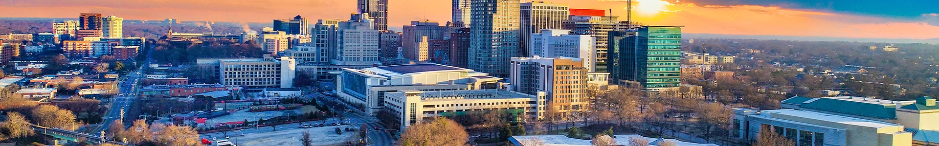 Panoramic skyline view of downtown Raleigh at sunset, featuring modern high‑rise buildings and tree‑lined neighborhoods.