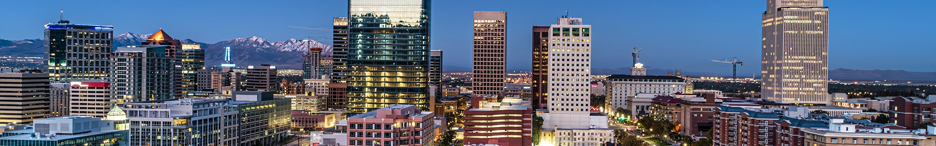 Panoramic view of downtown Salt Lake City at dusk, featuring illuminated skyscrapers, mountain backdrops, and vibrant city lights.
