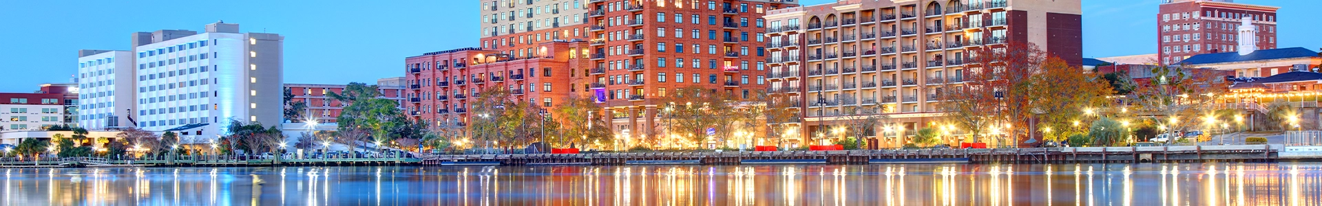 Panoramic waterfront view of downtown Wilmington at dusk, featuring illuminated buildings reflecting on the Cape Fear River.