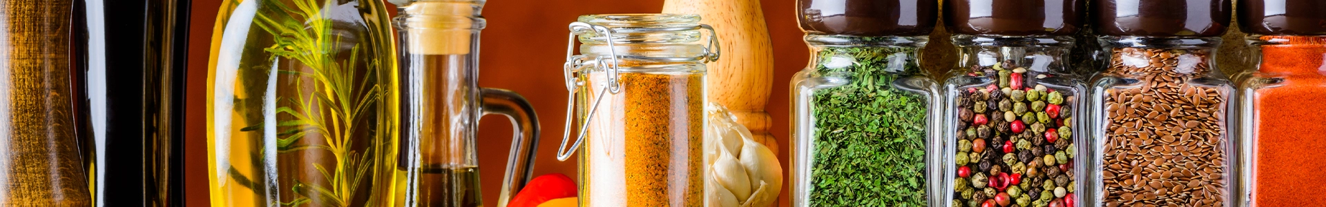 Rustic kitchen shelf displaying olive oil, infused herbs, jars of salt, peppercorns, dried spices, and colorful seasonings.
